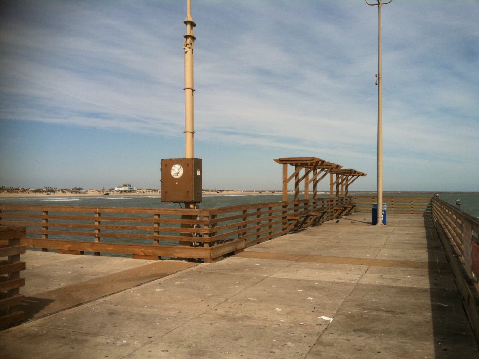 View along the wooden deck of Horace Caldwell Pier in Port Aransas, looking back toward shore — granite jetty channel and beach houses visible at the horizon, low rail covered shelters along the railing.