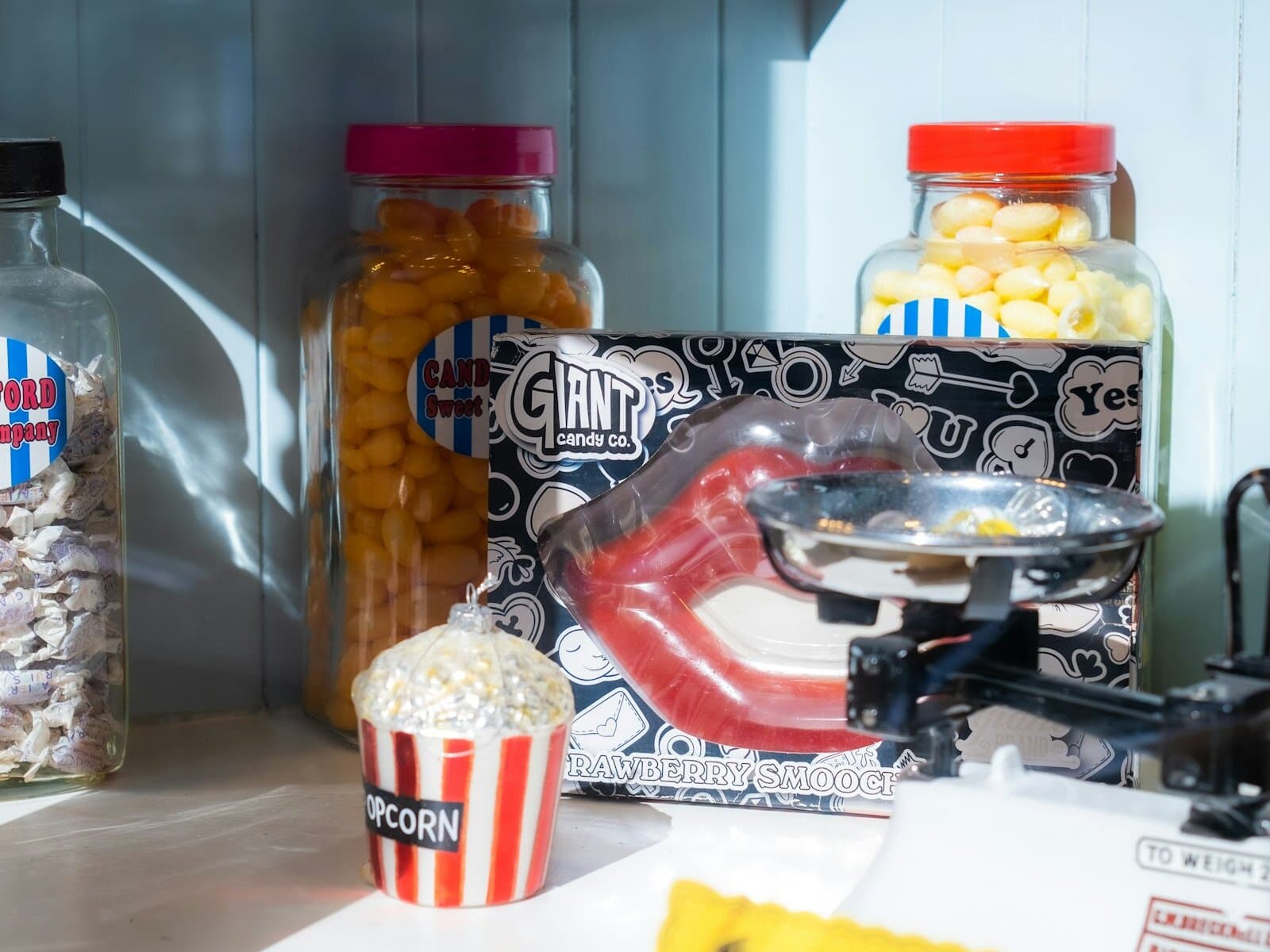 Vintage candy shop display with glass jars of colorful sweets, a Wonka chocolate bar prop and red-and-white popcorn boxes on a wooden counter — representative of the old-fashioned candy store experience at Winton's Island Candy.