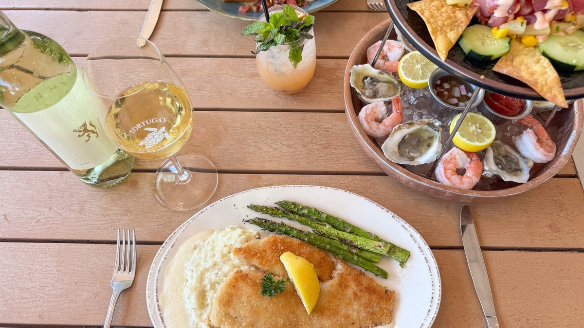 Table at Tortuga's Saltwater Grill set with panko-crusted fish over grits, a tiered seafood tower, and a wine glass etched with the Tortuga's turtle logo